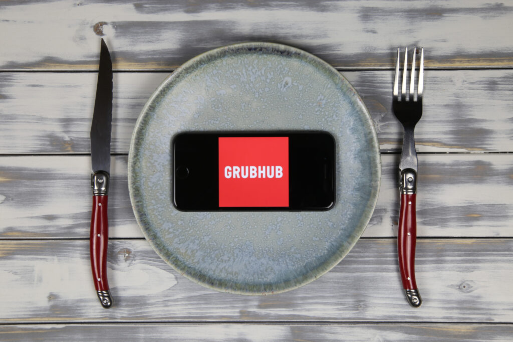 close up of mobile phone screen with logo lettering of food delivery service grubhub on wood table with dish and cutlery 205467998